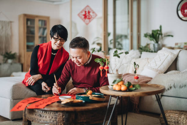 coppia senior asiatica che pratica la calligrafia cinese per il capodanno cinese fai chun (messaggi di buon auspicio) scrivendola su un pezzo di carta rossa - chinese heritage foto e immagini stock