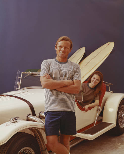 homem de pé em carro antigo, mulher no fundo - image created 1960s 1960s style beach women imagens e fotografias de stock