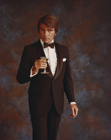 Young latin man poses in formal wear for a portrait. He is wearing a grey suit, blue tie and white shirt. He is serious. The background is black.