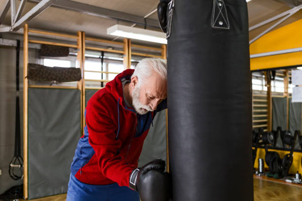 Senior in gym boxing taking a break leaning on a punching bag Senior man is tired from training boxing in gym leaning on a punching bag old man boxing stock pictures, royalty-free photos & images