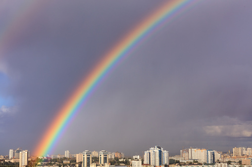 A bright large and dense rainbow in a gray cloudy sky above city houses after the last thunderstorm, image with copy space.