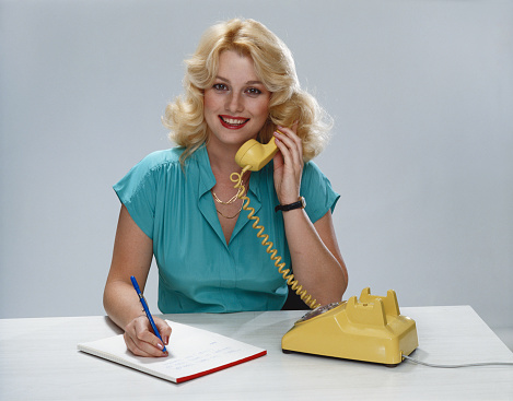 Filipino woman talking on landline phone call, using telephone with cord standing over yellow background. Cheerful model using office phone with cord having remote discussion in studio