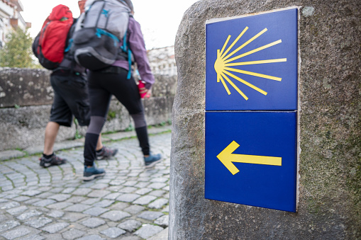Way of St. James sign and unfocused pilgrims at background. Yellow scallop sign pilgrimage to Santiago de Compostela
