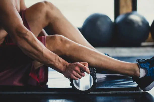 Muscular Caucasian man massaging his calves with a foam roller in an indoor gym.