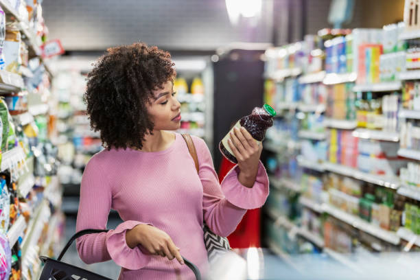 Young African-American woman shopping in supermarket A young African-American woman in her 20s shopping in a grocery store, carrying a shopping basket. She is reading the ingredient label on a bottle. nutrition label stock pictures, royalty-free photos & images