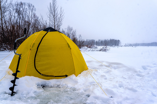 A winter yellow fishing tent stands next to an ice pack on snow and ice in a Blizzard on the lake near the trees.