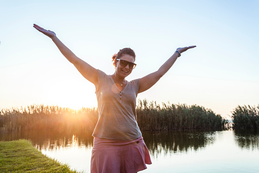 Adult woman raising her arms looking at camera in nature. The woman is looking at camera wearing sunglasses and backlit by sun at dawn. DSRL outdoors photo taken with Canon EOS 5D Mk II and Canon EF 17-40mm f/4L IS USM Wide Angle Zoom Lens