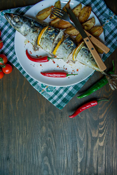 Baked mackerel with lemon and baked potatoes on a white plate. Fresh vegetables . Cherry, chili pepper. Cutlery. View from above. Wooden table. Space for text.