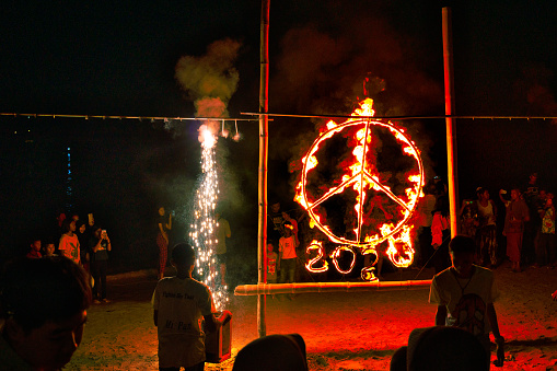 Koh Lanta, Krabi province, Thailand - December 31 2019:  The clock has just turned midnight and the New Year of 2020 just begun.  One large peace symbol saying 2020 has been set on fire at an open air beach party in Asia.  Crowds of young Thai people gather around to celebrate and live stream the event via mobile telephones.