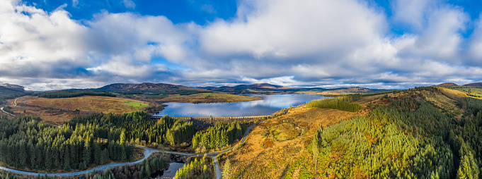 A panoramic aerial view, captured by a drone, of a  fresh water reservoir created by building a dam across a Scottish river. The dam was part of the Galloway hydro electric scheme which was built between 1930 and 1936. The loch is in part of the country popular for walking and mountain biking.\nThe panorama was created by merge several images together.