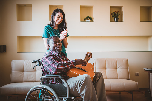 A female doctor is at an in-home visit of her older physically disabled patient of Afro-American ethnicity. A senior patient man of Afro-American ethnicity in wheelchairs is playing an acoustic guitar to a female healthcare worker in a green uniform.