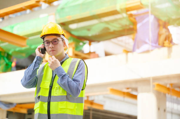 young asian engineers are working on the construction site. wear a yellow helmet safety and uniform staff. hand holding portable radio transceiver for communication - mobile work imagens e fotografias de stock