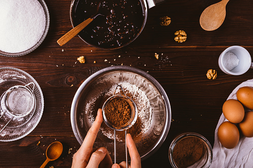 Top view woman's hands sift cocoa powder into bowl with flour to make dough for chocolate brownie cake on kitchen wooden table among ingredients.