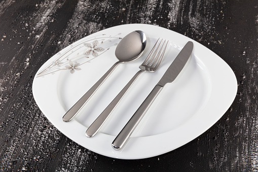 Empty plate, fork, knife, spoon, cutlery isolated on black wooden background, top view.