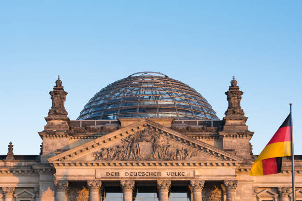 The historic parliament building rises against a cloudy night sky with the German national flag visible on the roof. Architectural elements are illuminated, emphasizing structure and symmetry.