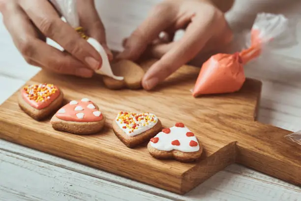 Photo of Decorating gingerbread cookies with icing. Woman hand decorate cookies in shape of heart, closeup