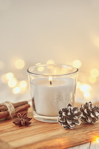 White burning candle with decorations and garland lights in bokeh on wooden table. Cozy home and hygge concept