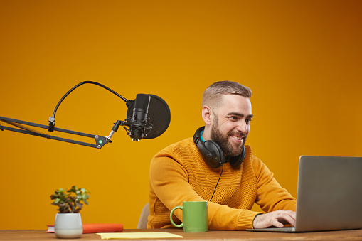 Horizontal medium studio shot of happy young Caucasian man typing something on his laptop, mustard background