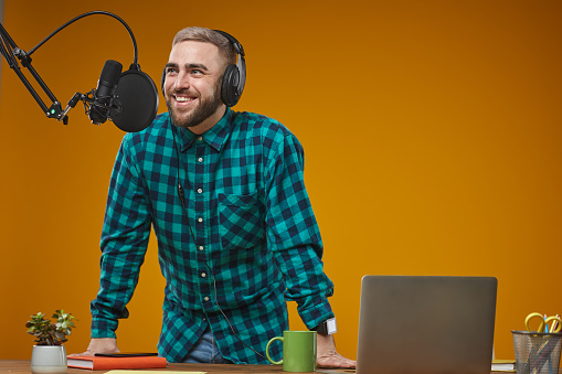 Horizontal studio shot of Caucasian young adult man wearing turquoise checked shirt recording radio content, mustard background