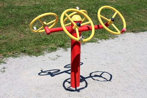 Tai chi spinner colorful red and yellow outdoor public park fitness exercising equipment with four rotary wheels on different heights surrounded with gravel and freshly cut grass in local public park