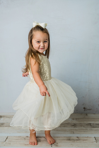 pretty preschool age girl wearing a gold sequined dress and a white bow spinning and twirling in a pale neutral room. She is happy and having fun playing.