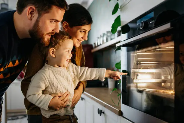 Young family with son is looking in the oven and waiting for their cookies which they prepared to be finished. They are smiling and happy. They are wearing warm wool sweaters.