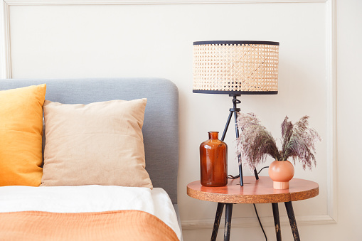 Stylish lamp on wooden nightstand next to flower in big glass vase in scandinavian bedroom interior.