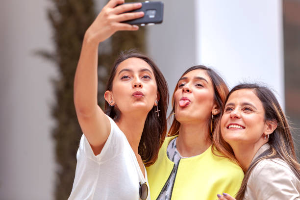 tres colegas latinoamericanas son vistos fuera de su edificio de oficinas en la luz del sol de la mañana: es hora de selfies para las redes sociales; se les ve posando para algunos medios de comunicación en línea - obviamente una parte de su vida milena - mobile phone group of people human face latin american and hispanic ethnicity fotografías e imágenes de stock