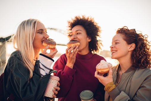 Three girls drinking coffee and eating donuts outdoors.