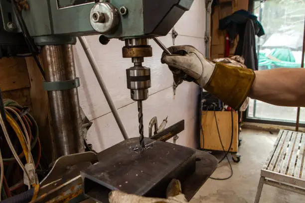 A close up view of a metalworker operating a bench drill inside his workshop. Heavy duty machine is used for drilling a hole through metal.
