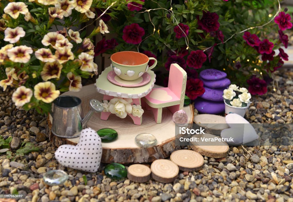 Tiny table, chair and cup by flowerpot with petunia flowers in the garden. Tiny table, chair and cup by flowerpot with petunia flowers in the garden. Lovely still life for greeting cards, wedding or birthday concept. Vintage summer background Fairy Stock Photo