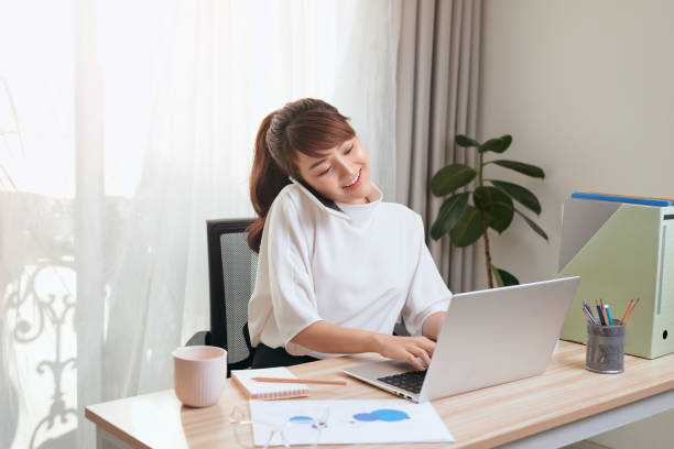 istock Young Asian woman using her cellphone while using laptop and working from home 1197066160