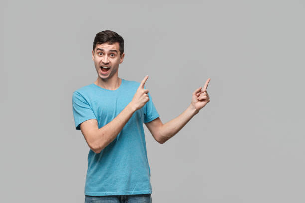 istock Pretty man in a blue tee pointing with a finger at the copy space isolater over grey background. 1197069614
