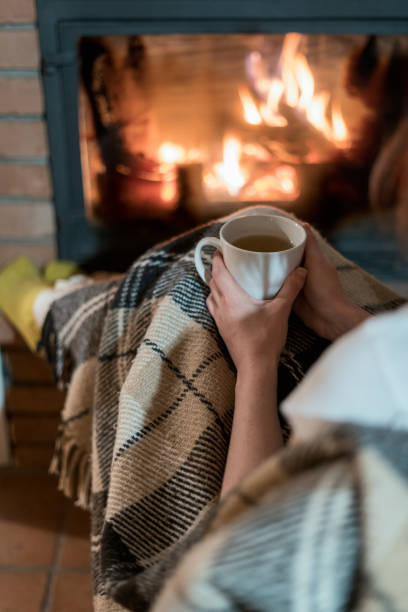 abgeschnittene ansicht der jungen erwachsenen frau sitzt mit tasse getränk in der nähe von kamin in komforthaus. mädchen verbringen wochenende tag zu hause - blanket fireplace winter women stock-fotos und bilder