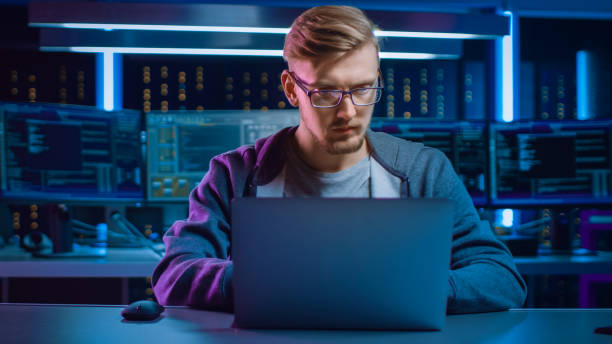 istock Portrait of Software Developer / Hacker / Gamer Wearing Glasses and Headset Sitting at His Desk and Working / Playing on Laptop. In the Background Dark High Tech Environment with Multiple Displays. 1197070963