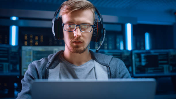 istock Portrait of Software Developer / Hacker / Gamer Wearing Glasses and Headset Sitting at His Desk and Working / Playing on Laptop. In Background Dark Neon High Tech Environment with Multiple Displays. 1197070983