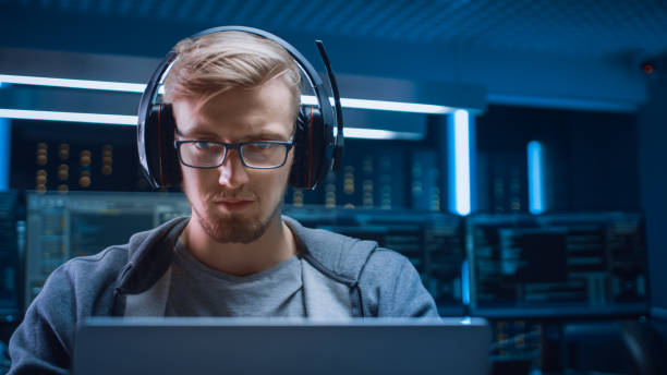 istock Portrait of Software Developer / Hacker / Gamer Wearing Glasses and Headset Sitting at His Desk and Working / Playing on Laptop. In Background Dark Neon High Tech Environment with Multiple Displays. 1197070995