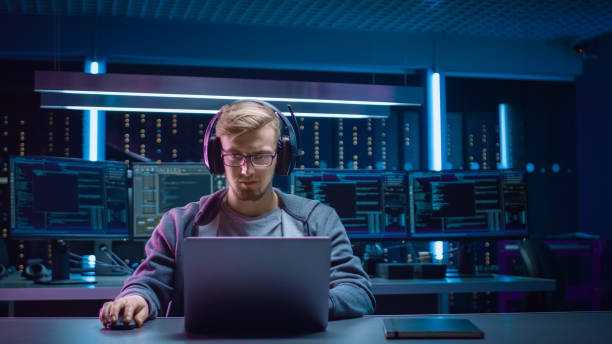 istock Portrait of Software Developer / Hacker / Gamer Wearing Glasses and Headset Sitting at His Desk and Working / Playing on Laptop. In Background Dark Neon High Tech Environment with Multiple Displays. 1197071020