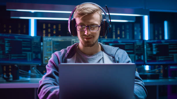 porträt von software-entwickler / hacker / gamer tragen brille und headset sitzen datiert an seinem schreibtisch und arbeiten / spielen auf laptop. im hintergrund dark high tech umgebung mit mehreren displays. - uncool stock-fotos und bilder