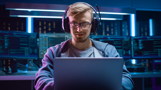 Retrato de desarrollador de software / Hacker / Gamer usando gafas y auriculares sentados en su escritorio y trabajando / jugando en el ordenador portátil. En el entorno de alta tecnología oscuro de fondo con varias pantallas. photo