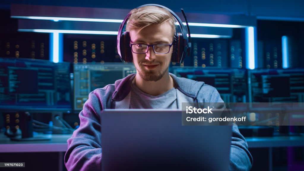 Portrait of Software Developer / Hacker / Gamer Wearing Glasses and Headset Sitting at His Desk and Working / Playing on Laptop. In the Background Dark High Tech Environment with Multiple Displays. Computer Programmer Stock Photo