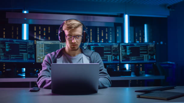 istock Portrait of Software Developer / Hacker / Gamer Wearing Glasses and Headset Sitting at His Desk and Working / Playing on Laptop. In Background Dark Neon High Tech Environment with Multiple Displays. 1197071029