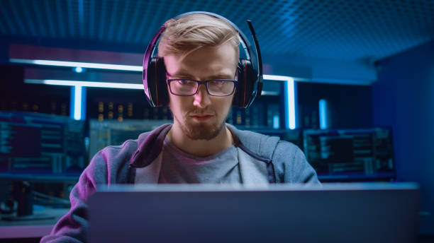 istock Portrait of Software Developer / Hacker / Gamer Wearing Glasses and Headset Sitting at His Desk and Working / Playing on Laptop. In Background Dark Neon High Tech Environment with Multiple Displays. 1197071058