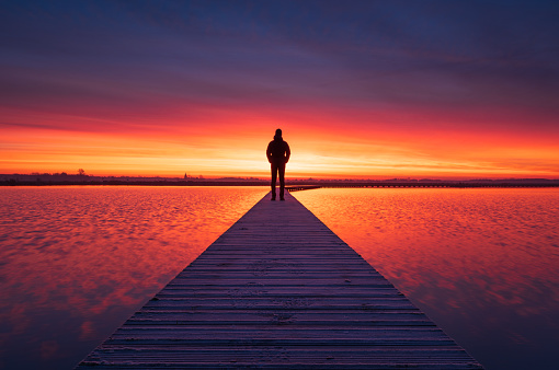 A man enjoying a colourful dawn on a small boardwalk over a lake.
