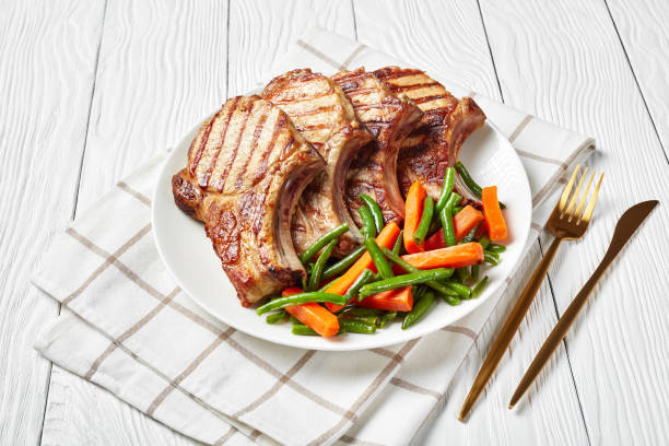 Grilled pork loin cutlets served with green bean and carrots salad Grilled pork loin chops on bones served with green bean and carrot salad on a white plate on a wooden table with golden fork and knife, horizontal view from above meat chop stock pictures, royalty-free photos & images
