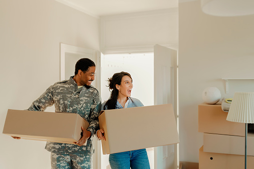 Happy wife and her african american army veteran husband in camouflage clothing with cardboard boxes in new house at moving day. First time home buyers.
