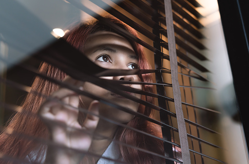 Asian woman looking through window blinds spying on neighbours - Young lonely millennial woman peeping through glass observing gossip and action outdoors - introvert, spy and intrusive concepts