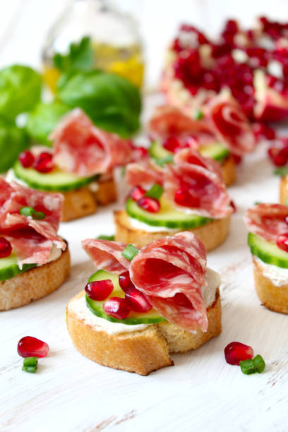 Canape or crostini with toasted baguette, light cheese, cucumber, pomegranate and salami on light background. stock photo