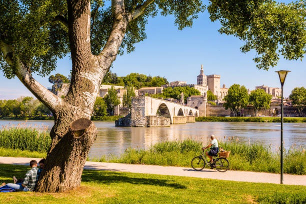 The banks of the Rhone on a sunny day, opposite the Avignon bridge and the Papal palace in Avignon, France. Avignon, France - May 16, 2018: People are biking or resting by a sunny day on the banks of the Rhone, opposite the Saint-Benezet bridge, also known as Avignon bridge, and the Papal palace. avignon france stock pictures, royalty-free photos & images