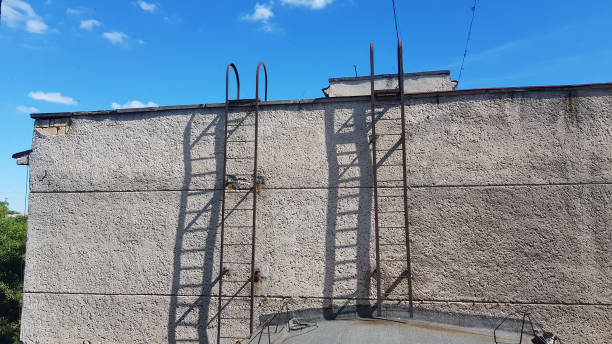 istock Cement weathered wall of old building on clear blue sky background with white clouds. Rooftop and two rusty metal ladders of fire exit. Textured wall surface and striped shadows from ladder. 1197149154